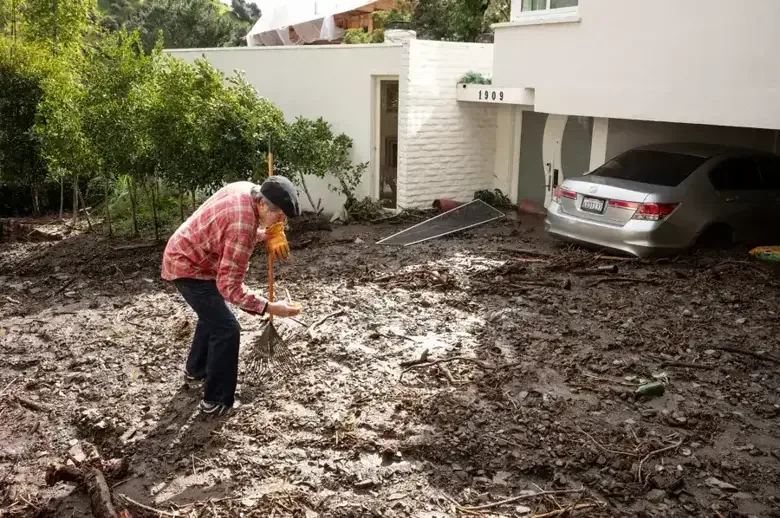 A person shovels storm mud beside a parked car and garage
